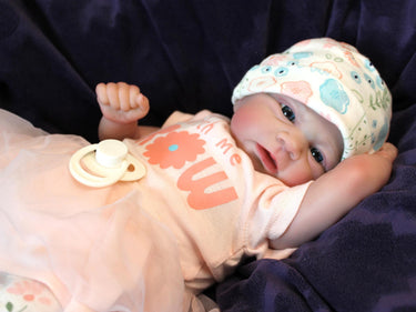A baby doll dressed in a pink shirt and wearing a hat, sitting against a soft background.